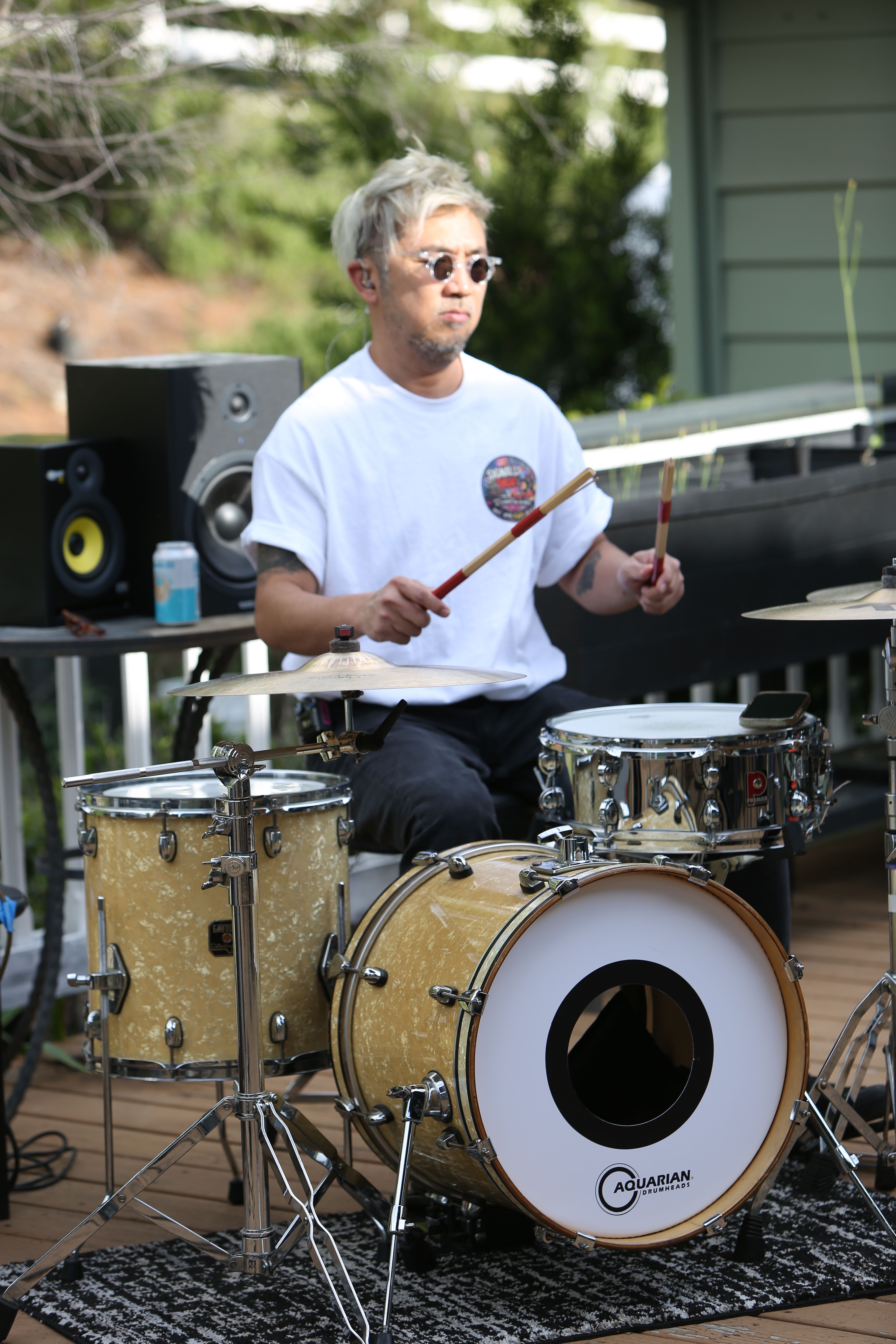 Woman playing drums outdoors with trees and a building in the background.