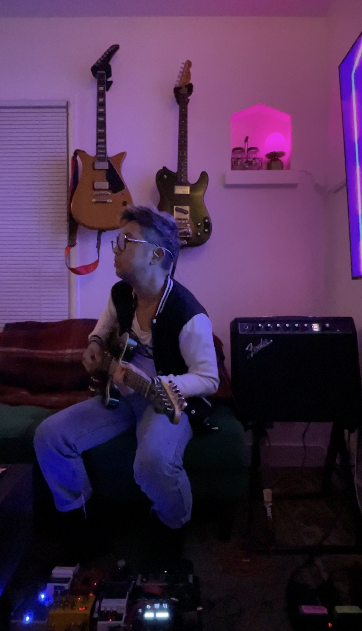 Person sitting on a couch playing guitar, with two guitars hanging on the wall behind, and a pink light on the wall.