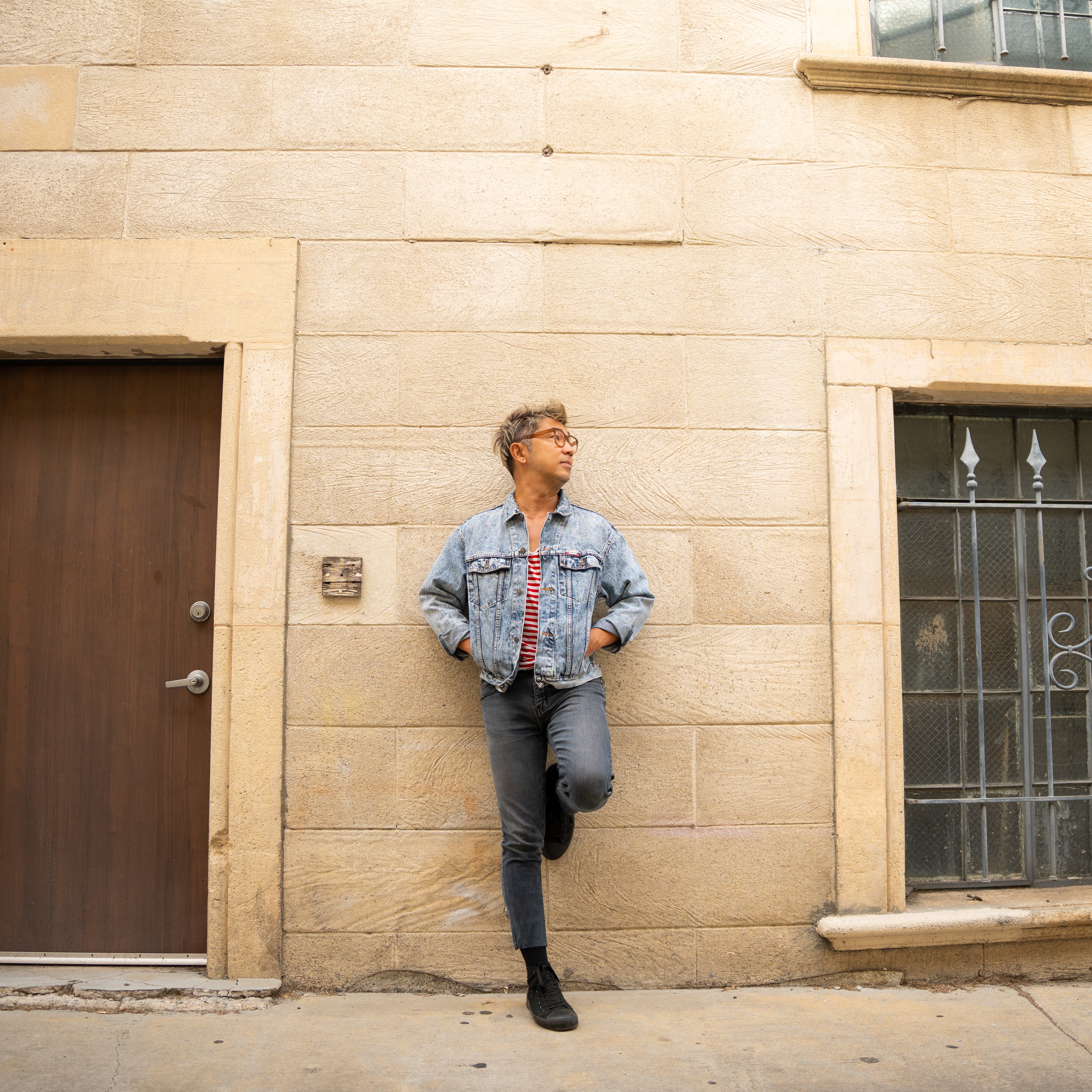 Person standing against a beige brick wall, wearing a denim jacket, jeans, and glasses, looking to the side.