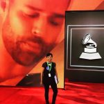 Person standing on red carpet in front of large screen with a record player icon, large face image in background.