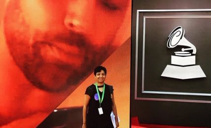 Person standing on red carpet in front of large screen with a record player icon, large face image in background.
