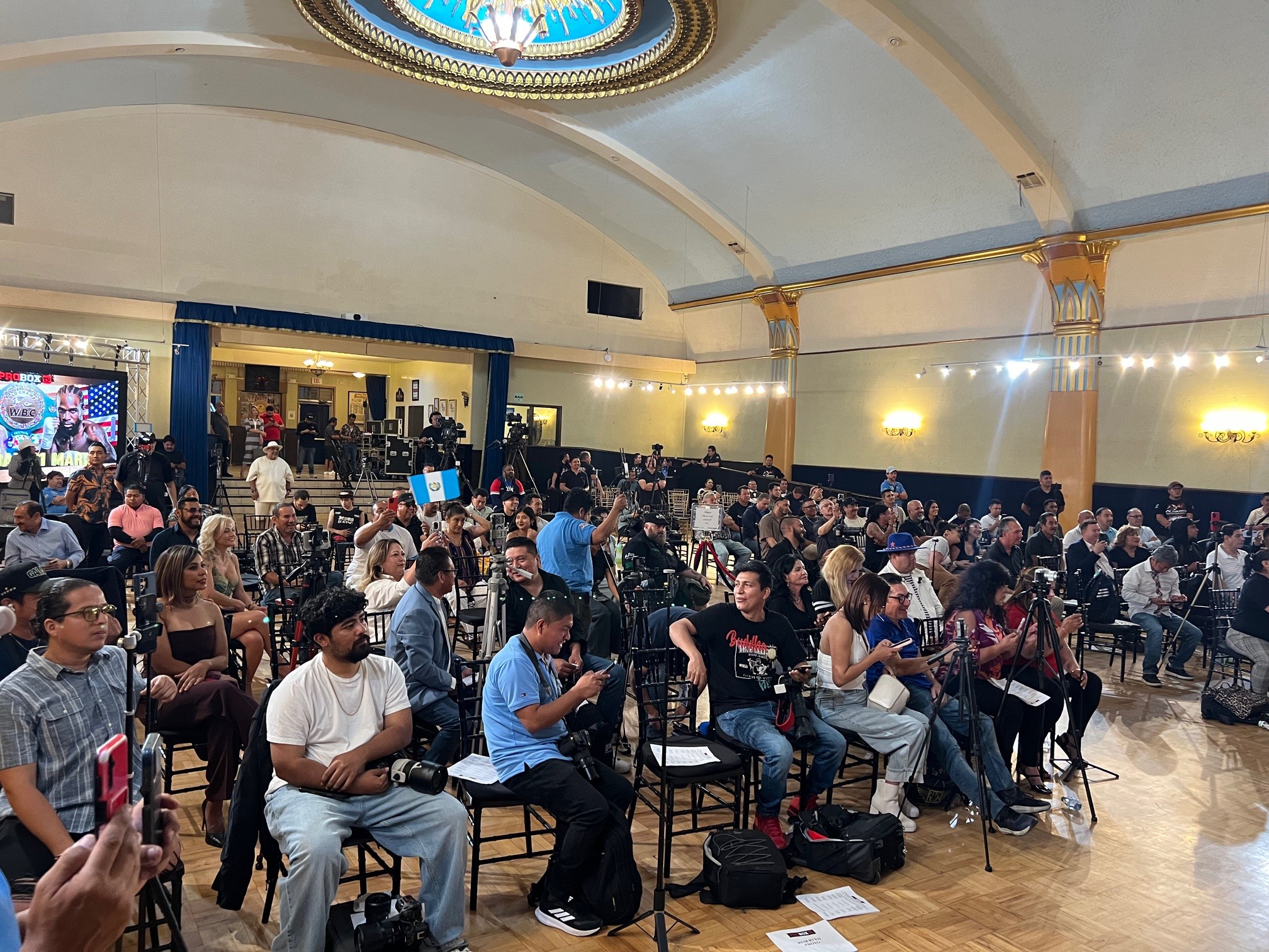 Large indoor event with many seated and standing people in a spacious hall with a high ceiling and decorative lighting.