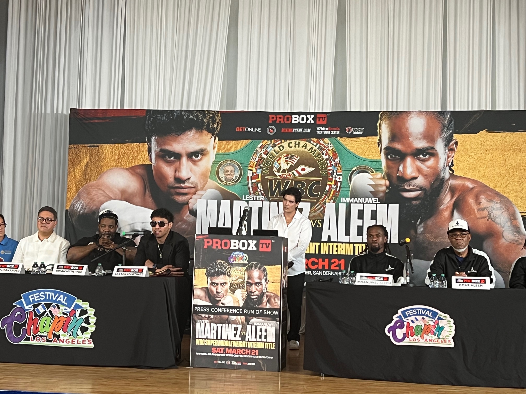 Press conference with five people seated at a table, promotional poster behind them, and a colorful banner in front.