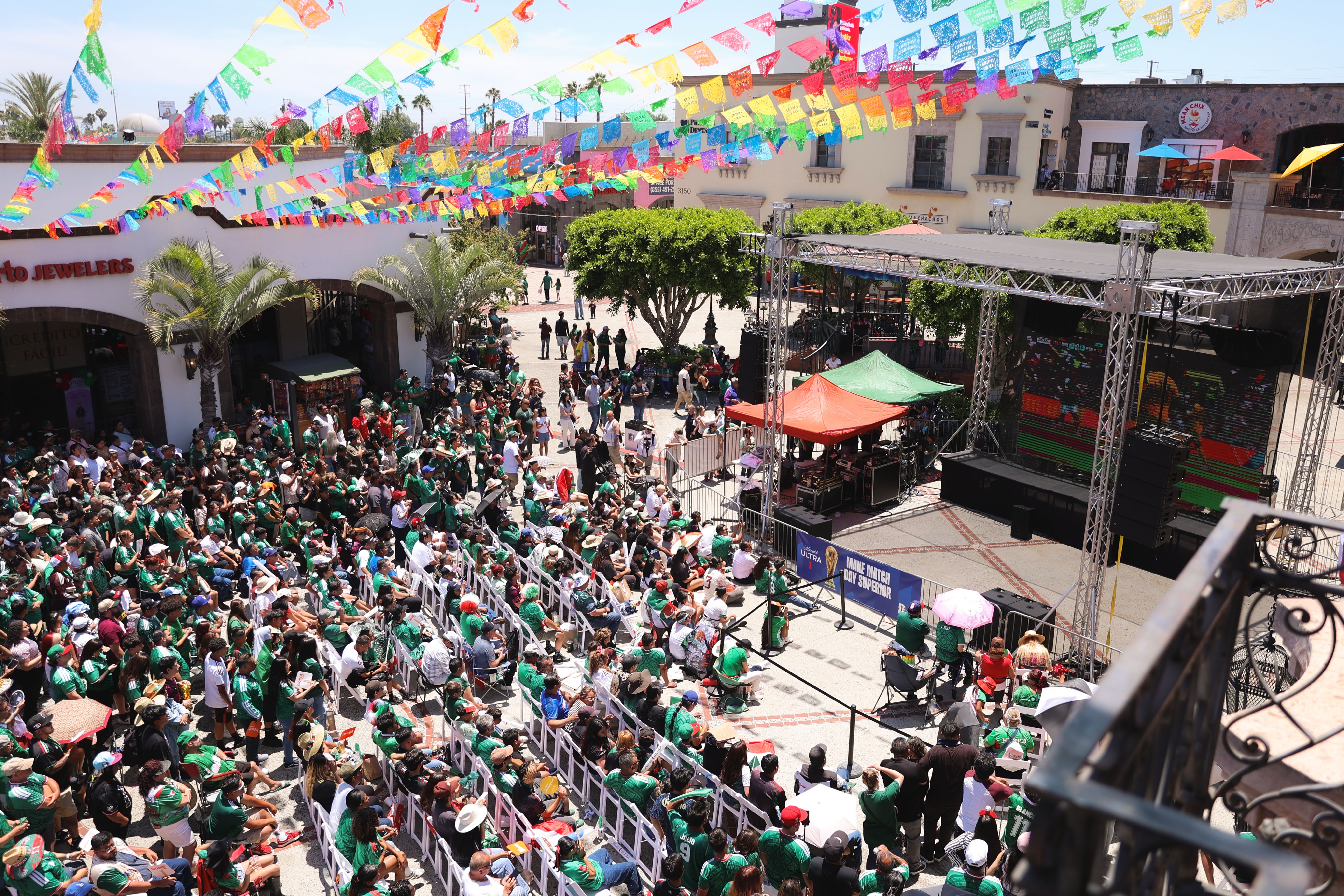 Large outdoor gathering with a stage, colorful banners, and many people attending an event in a town square.
