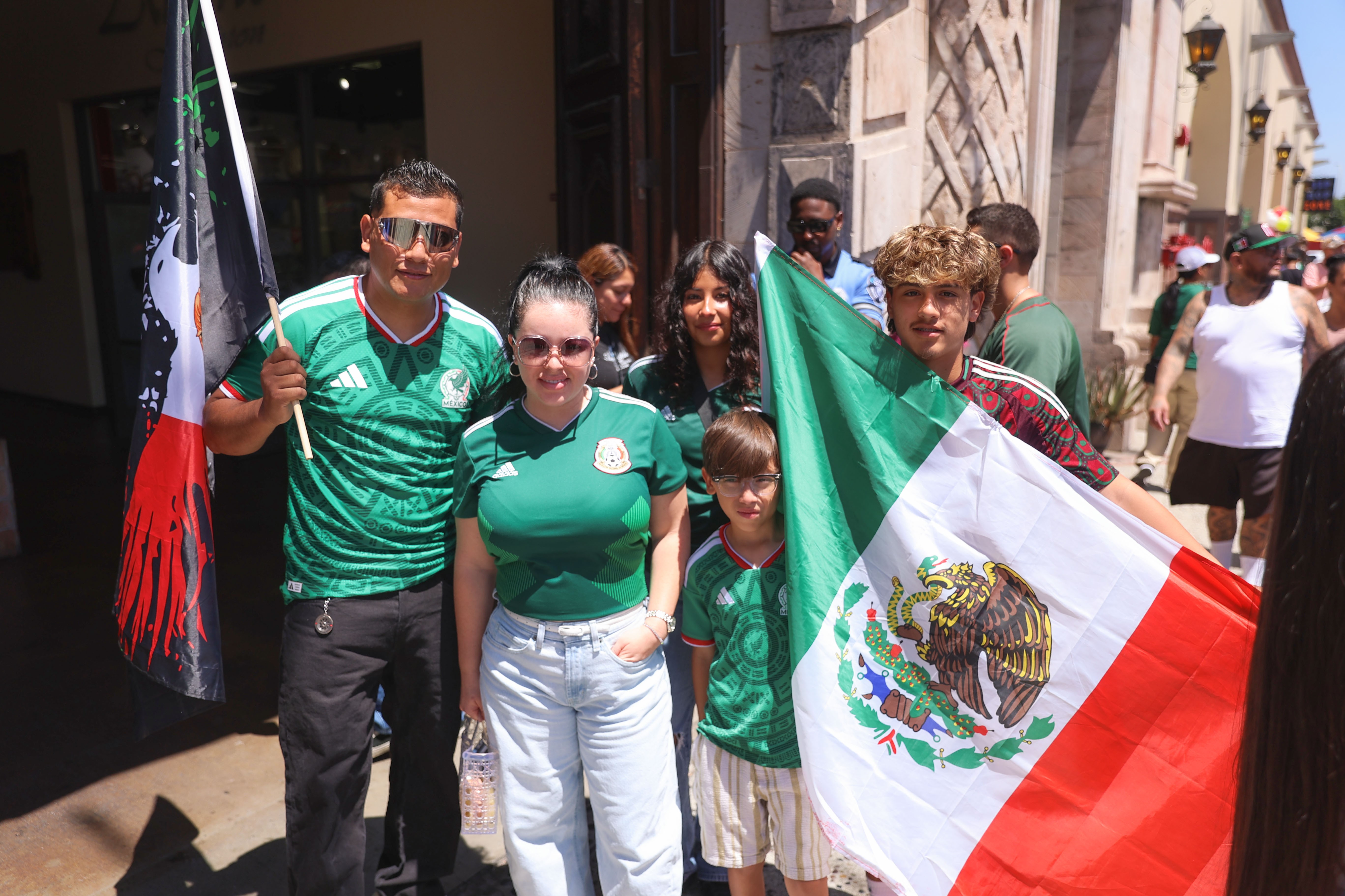 Group of people wearing green shirts holding Mexican flags outside a building, sunny day, some people in background.