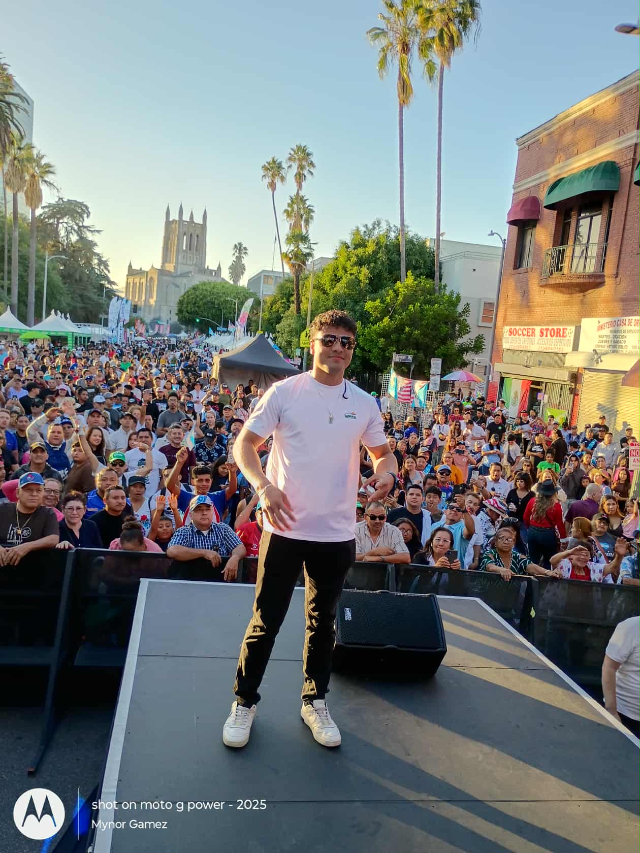 Young man standing on stage in front of large crowd outdoors with palm trees and buildings in background.