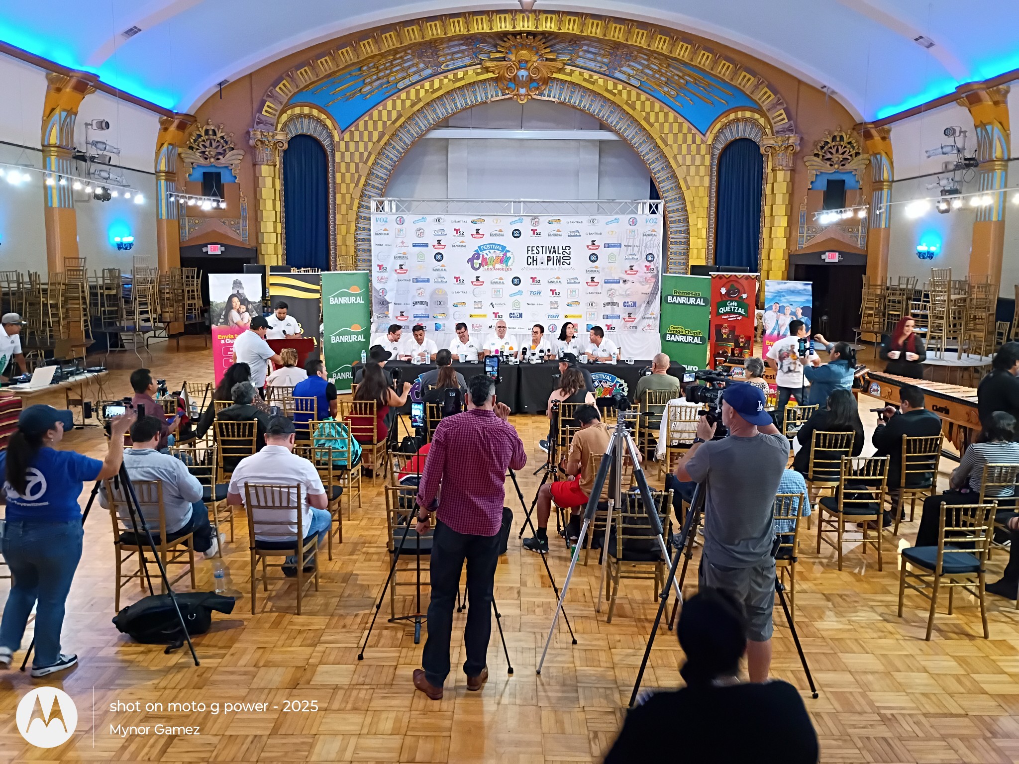 Event in a large hall with a stage, audience seated, and cameras recording, decorated with banners and screens.