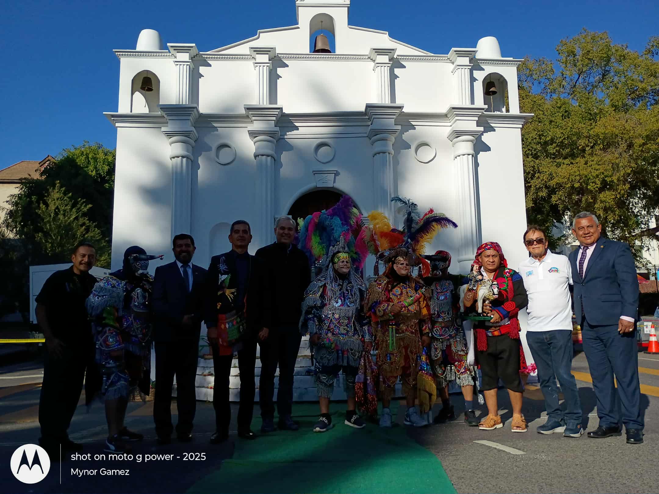 Group of people standing in front of a white building with arches and decorative elements, under a clear blue sky.