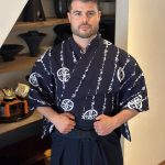 Man in traditional Japanese attire standing indoors with shelves of bowls behind him.