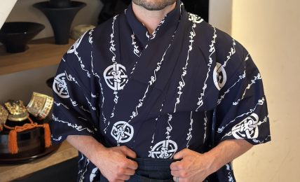 Man in traditional Japanese attire standing indoors with shelves of bowls behind him.