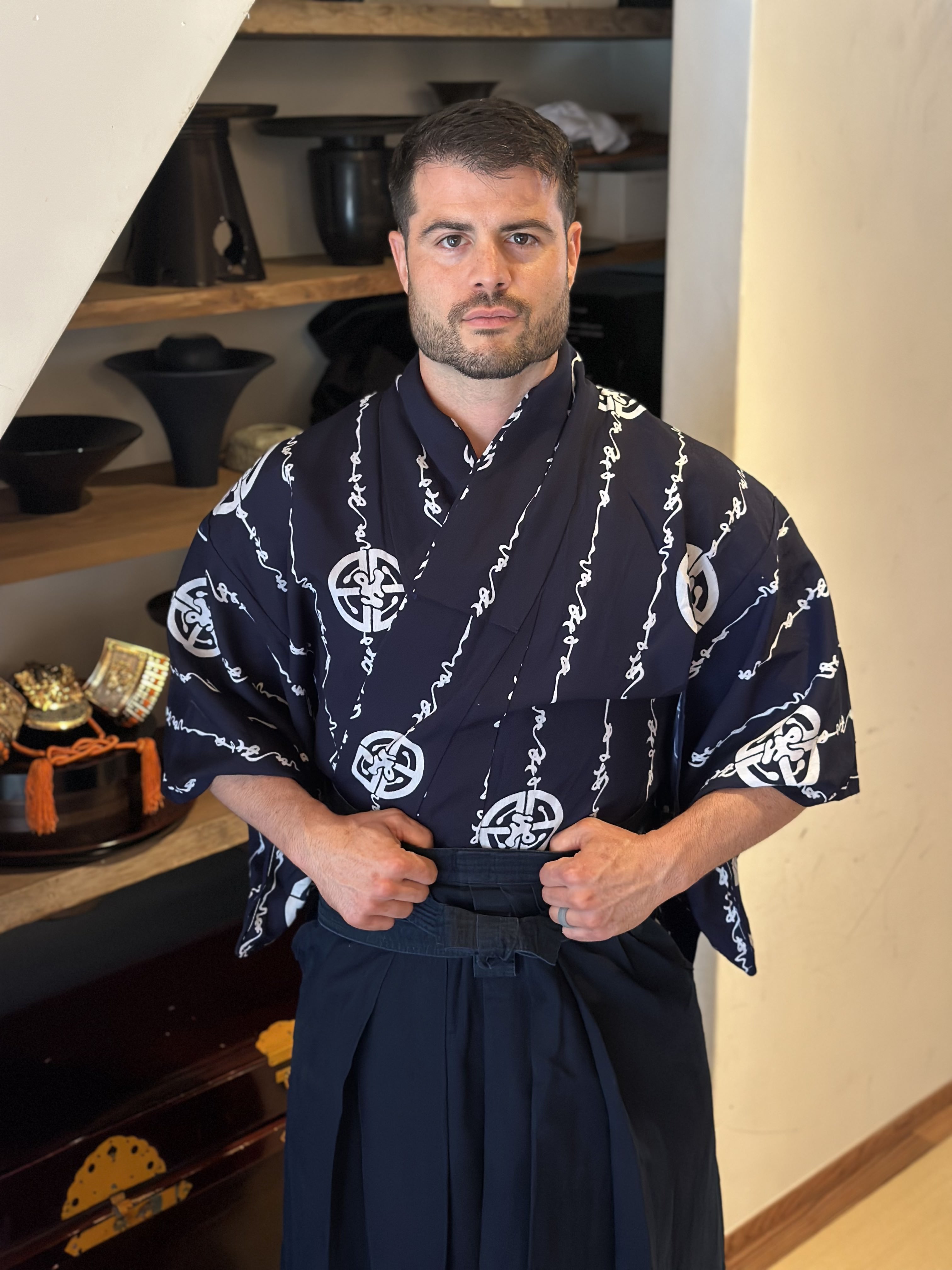 Man in traditional Japanese attire standing indoors with shelves of bowls behind him.