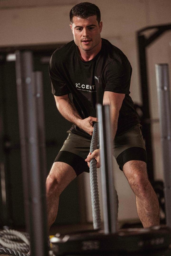 Young man in black shirt and shorts pulling a rope in a gym, focused expression, gym equipment in background.