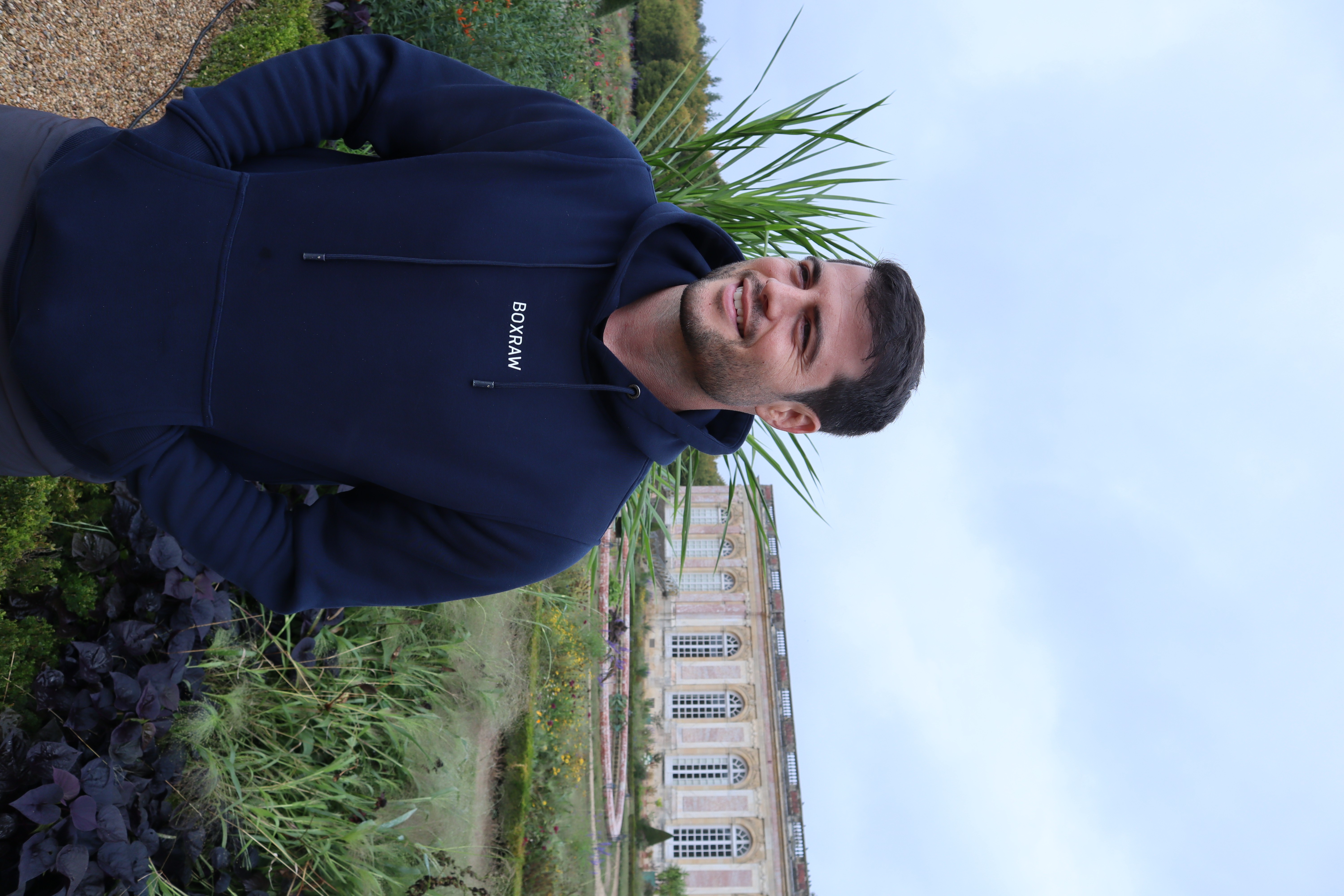 Man standing outdoors with plants and a building in the background, wearing a dark hoodie, smiling.