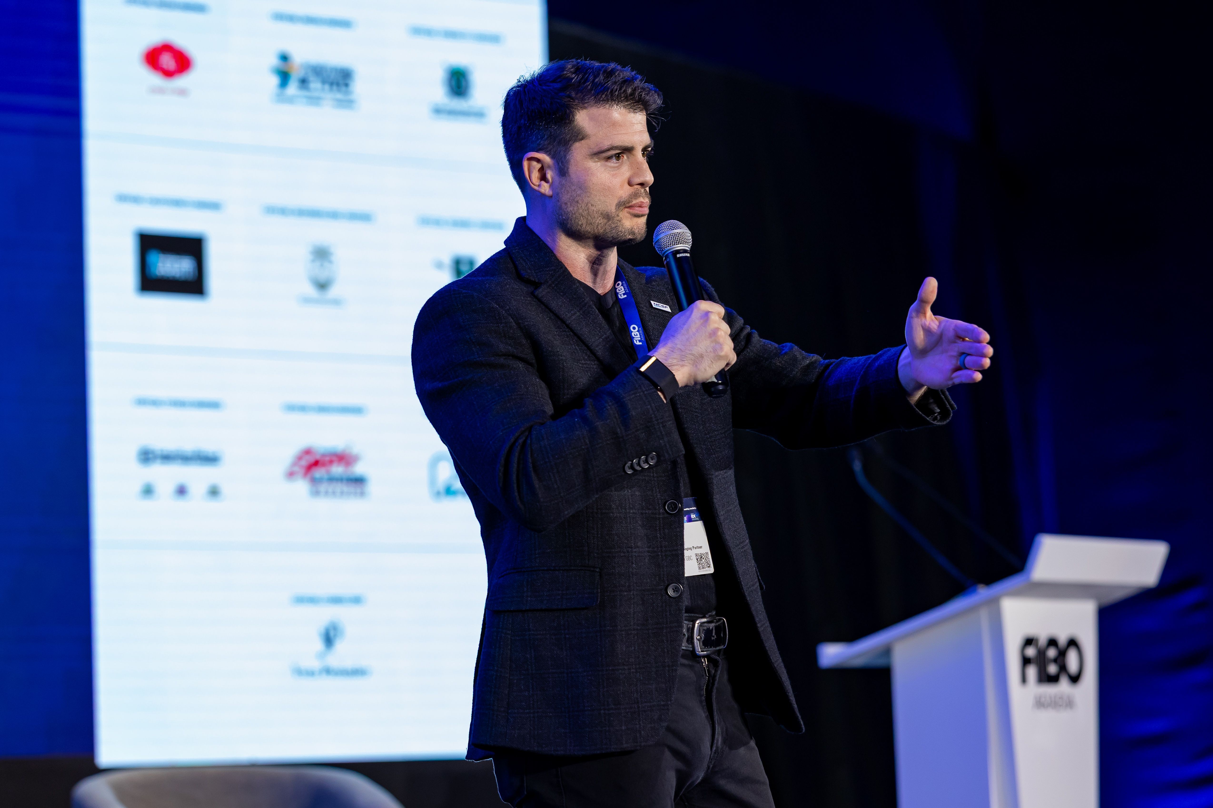 Man in dark suit speaking into a microphone on stage with a presentation screen behind him, gesturing with his right hand.