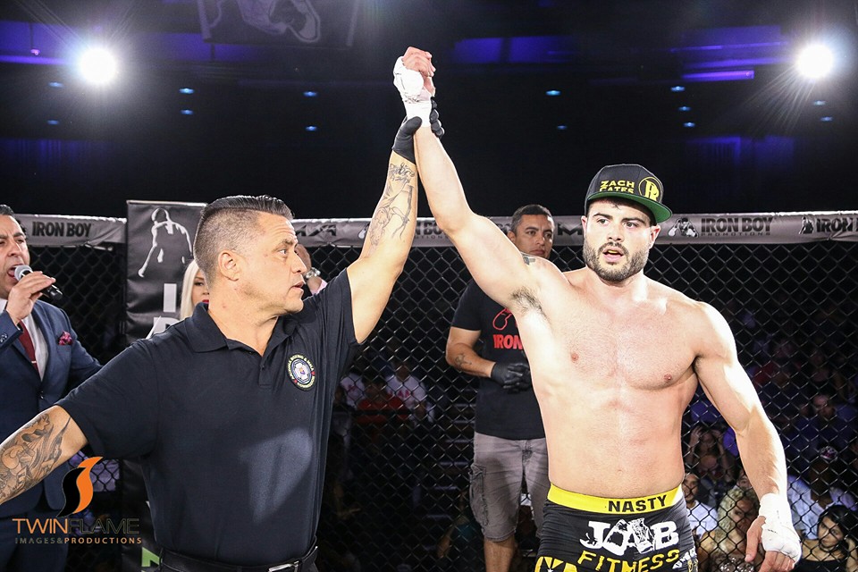 Mixed martial arts fighter with a cap raises his arm in victory inside the cage, referee confirms the win, audience in background.