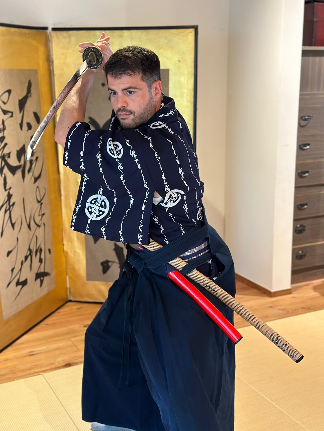 Man in traditional clothing holding a wooden staff, standing indoors near a folding screen and a cabinet.