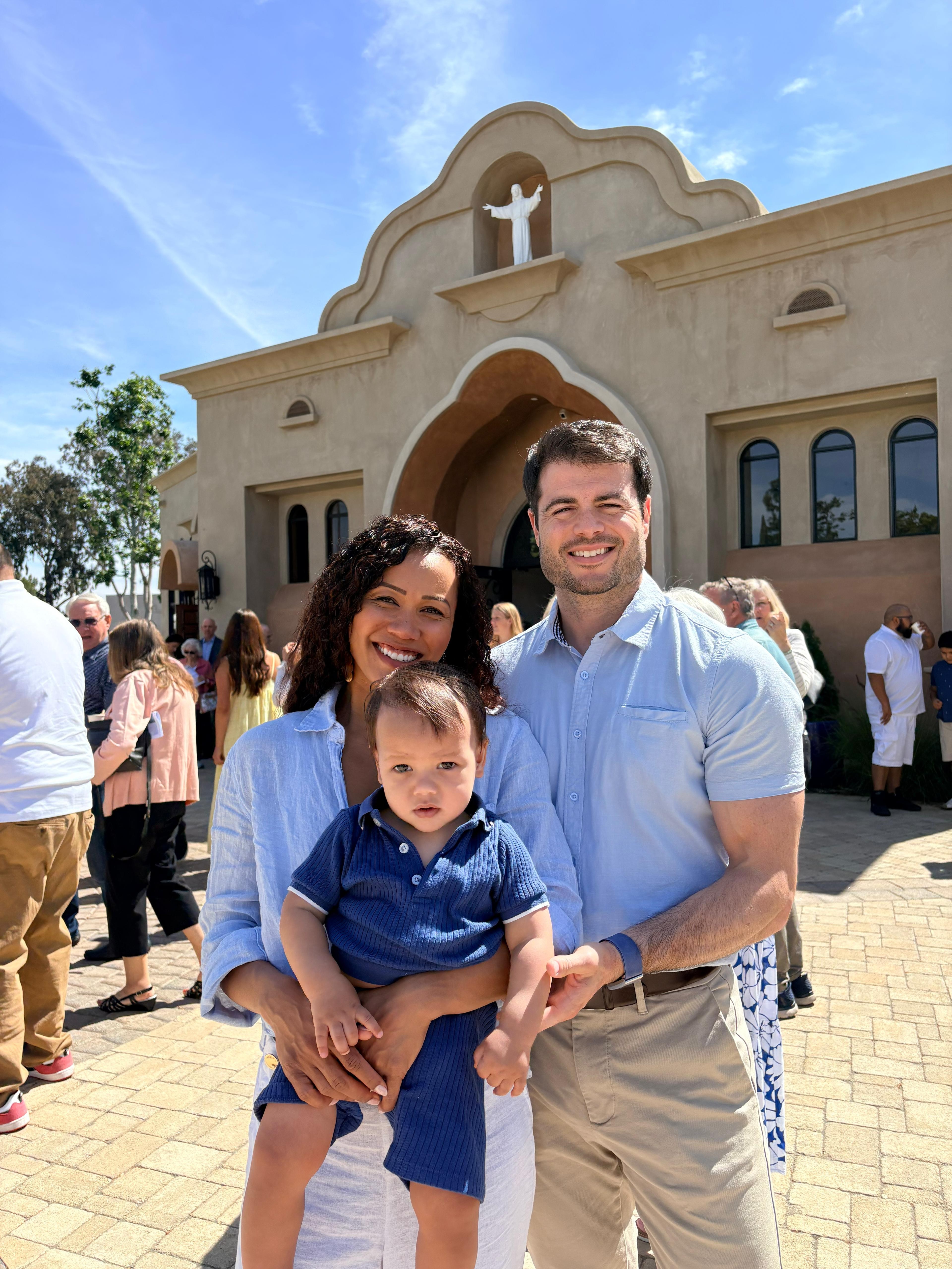 Family of three smiling outside a church, with people in the background, sunny day, blue sky.