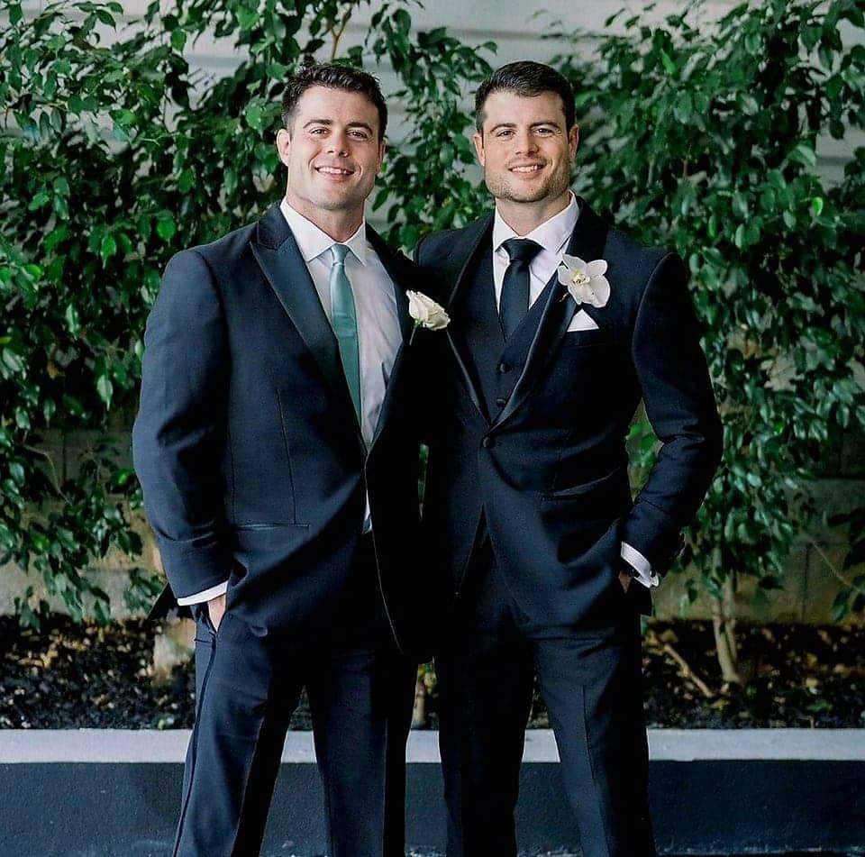 Two men in suits standing outdoors with greenery behind them, smiling, one with a white flower boutonniere.