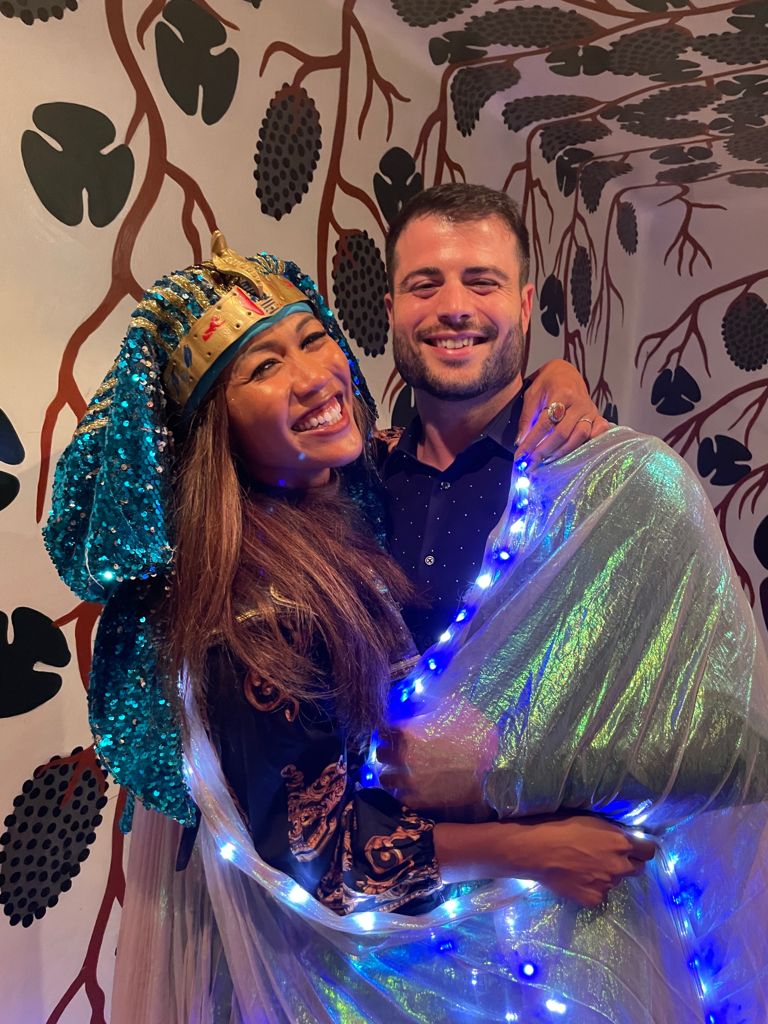 Two people smiling, one wearing a colorful headdress, the other holding a shiny fabric with blue lights, standing in front of a patterned wall.