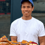 Smiling man in white t-shirt and blue cap holding a tray of assorted donuts outdoors.