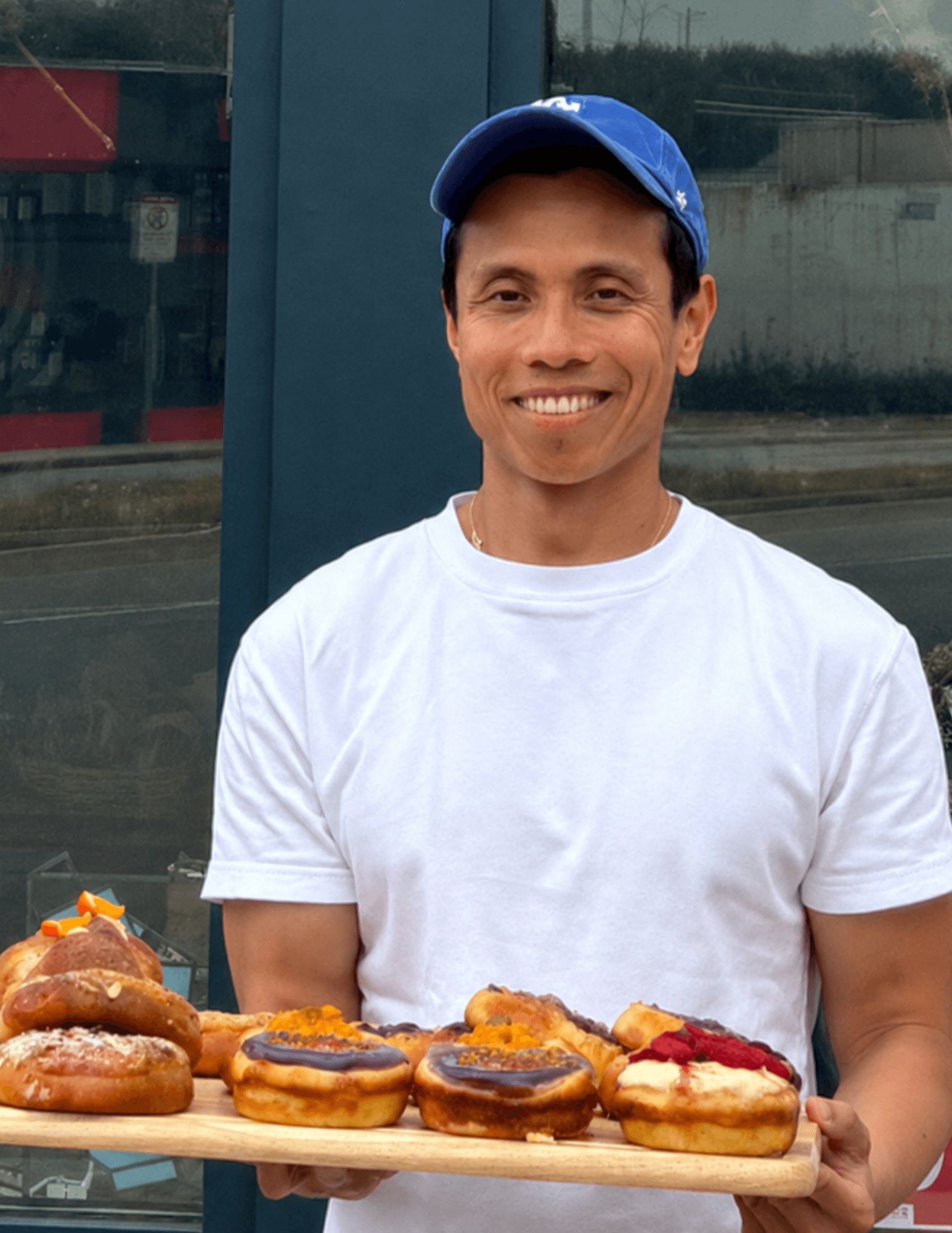 Smiling man in white t-shirt and blue cap holding a tray of assorted donuts outdoors.