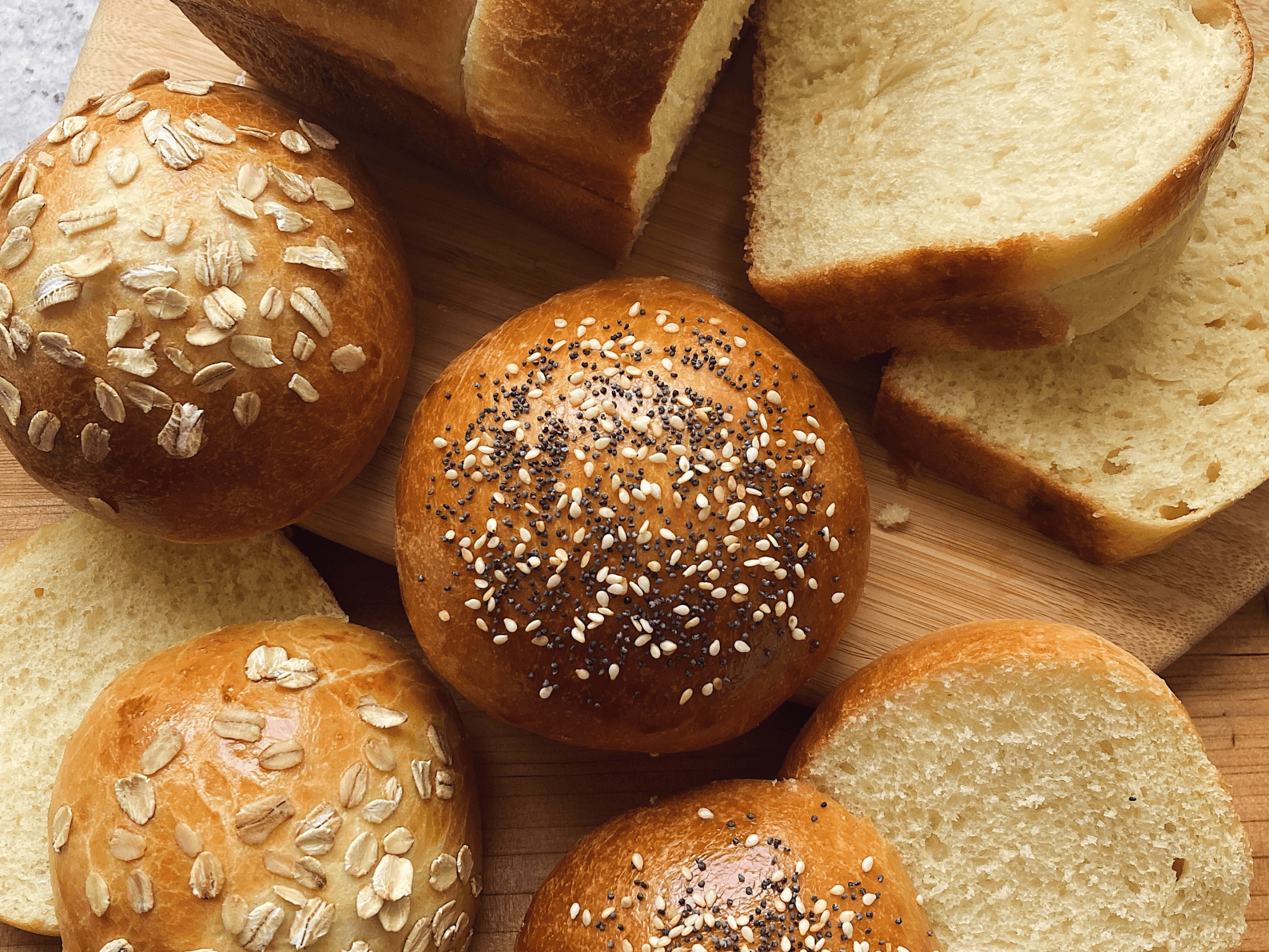 Assorted bread rolls and sliced bread on a wooden surface, some topped with oats and seeds.