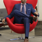 Man in a blue suit sitting in a red modern chair with a high back, in an office setting.