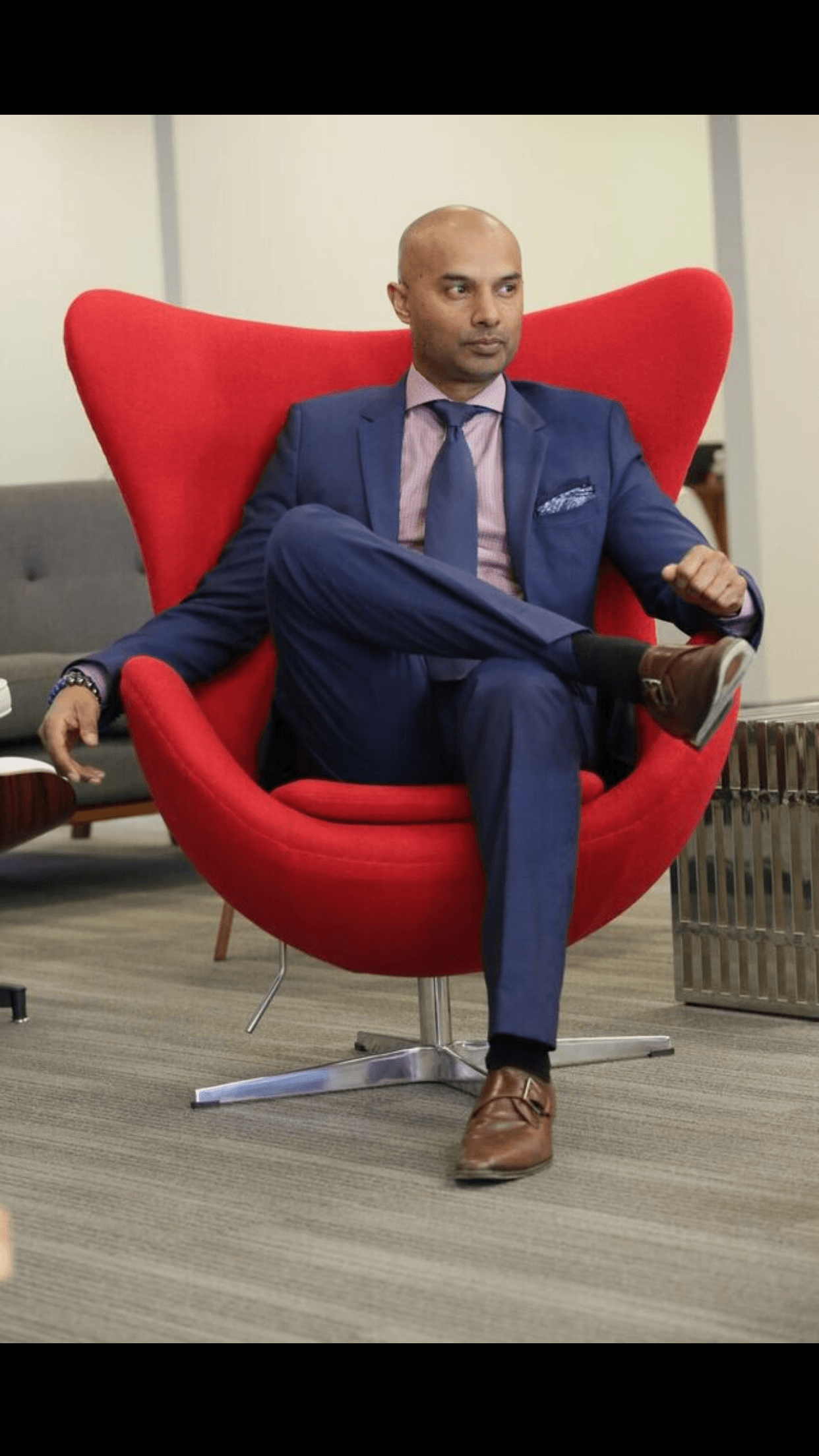 Man in a blue suit sitting in a red modern chair with a high back, in an office setting.