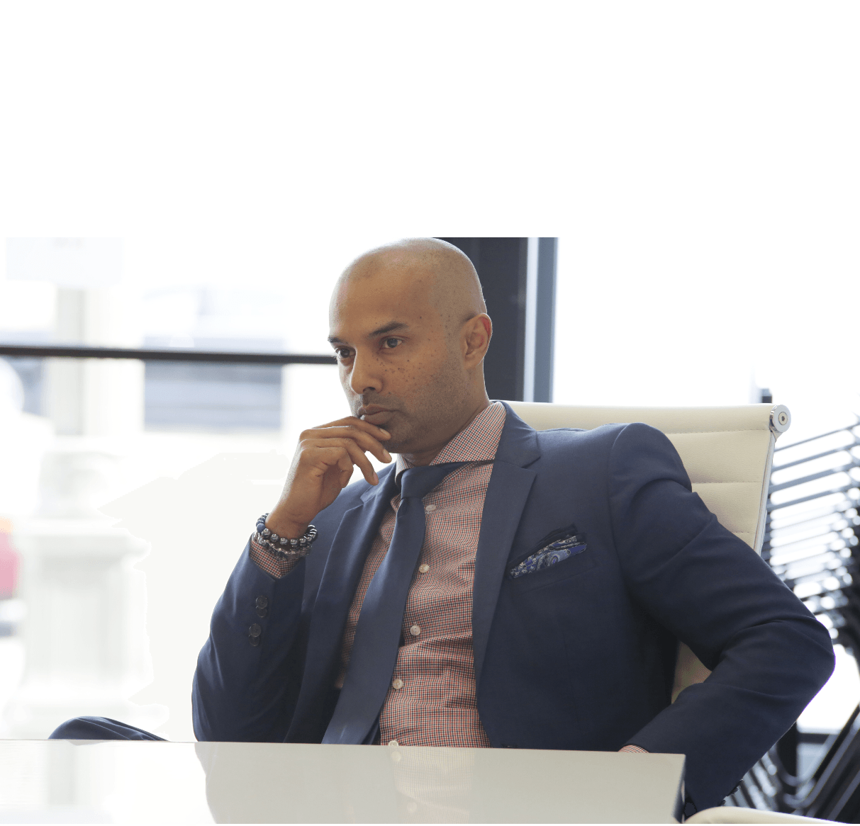 Man in suit sitting at desk, thinking, in an office with large windows behind him.