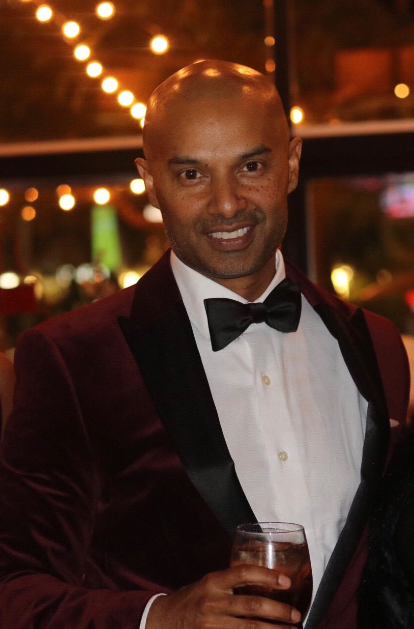 Man in tuxedo with bow tie holding a glass, smiling at an event with string lights in background.