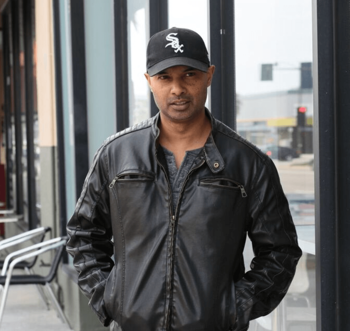 Man wearing a black leather jacket and a black baseball cap standing indoors near glass windows.