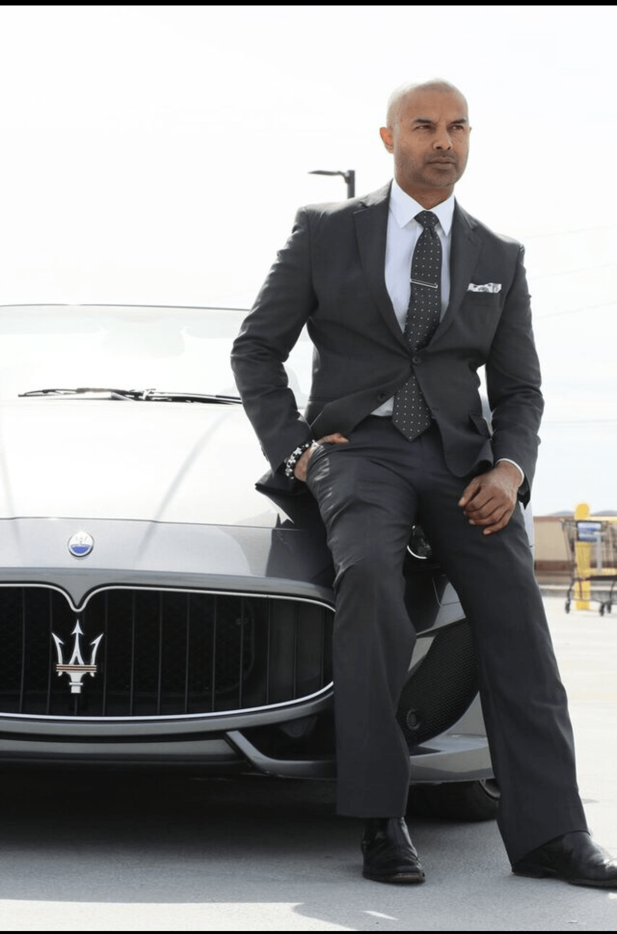 Man in suit leaning against a white Maserati car on a rooftop parking lot, with a cityscape in the background.