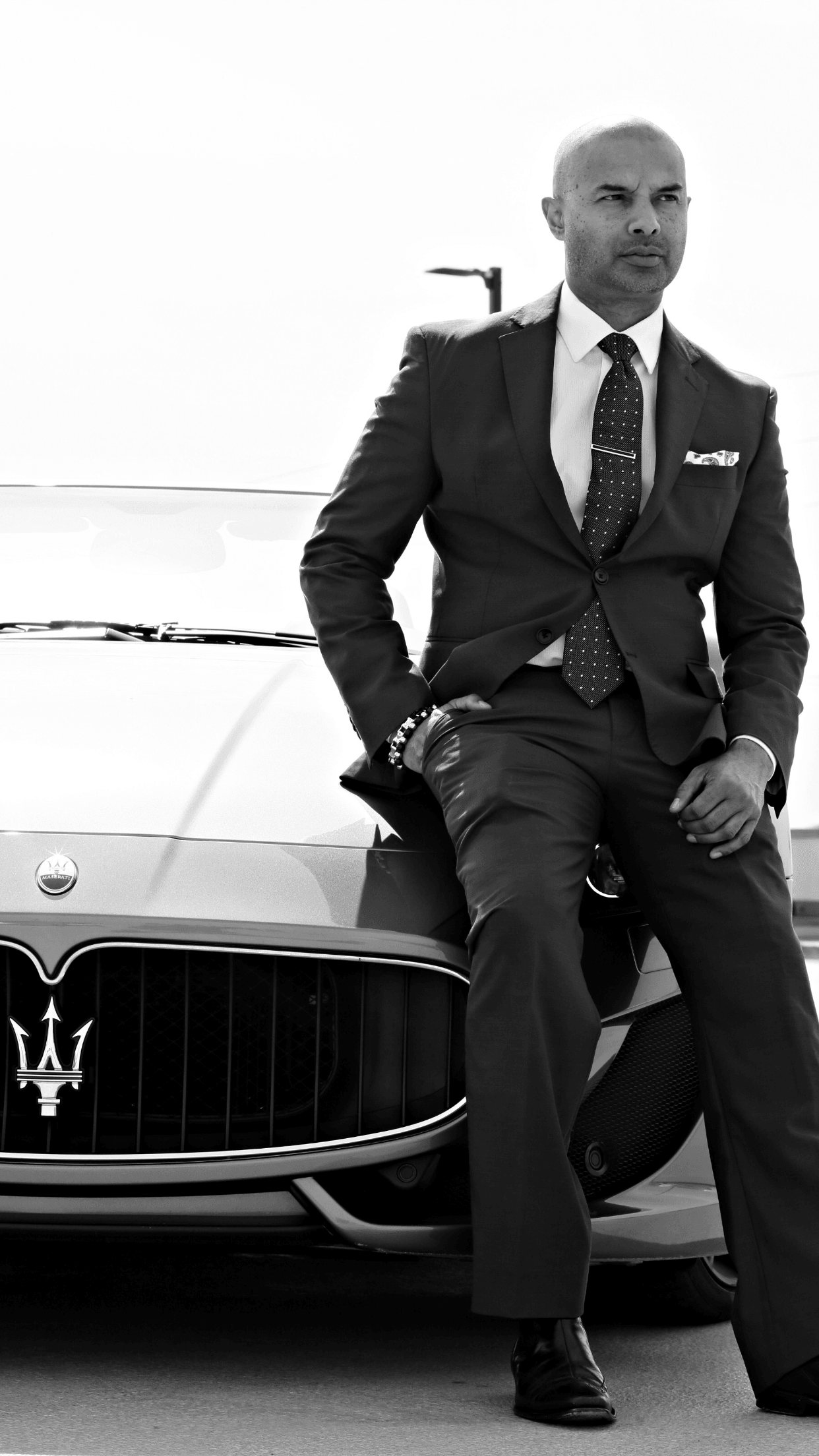 Man in suit leaning against a luxury car, looking to the side, with a plain background.