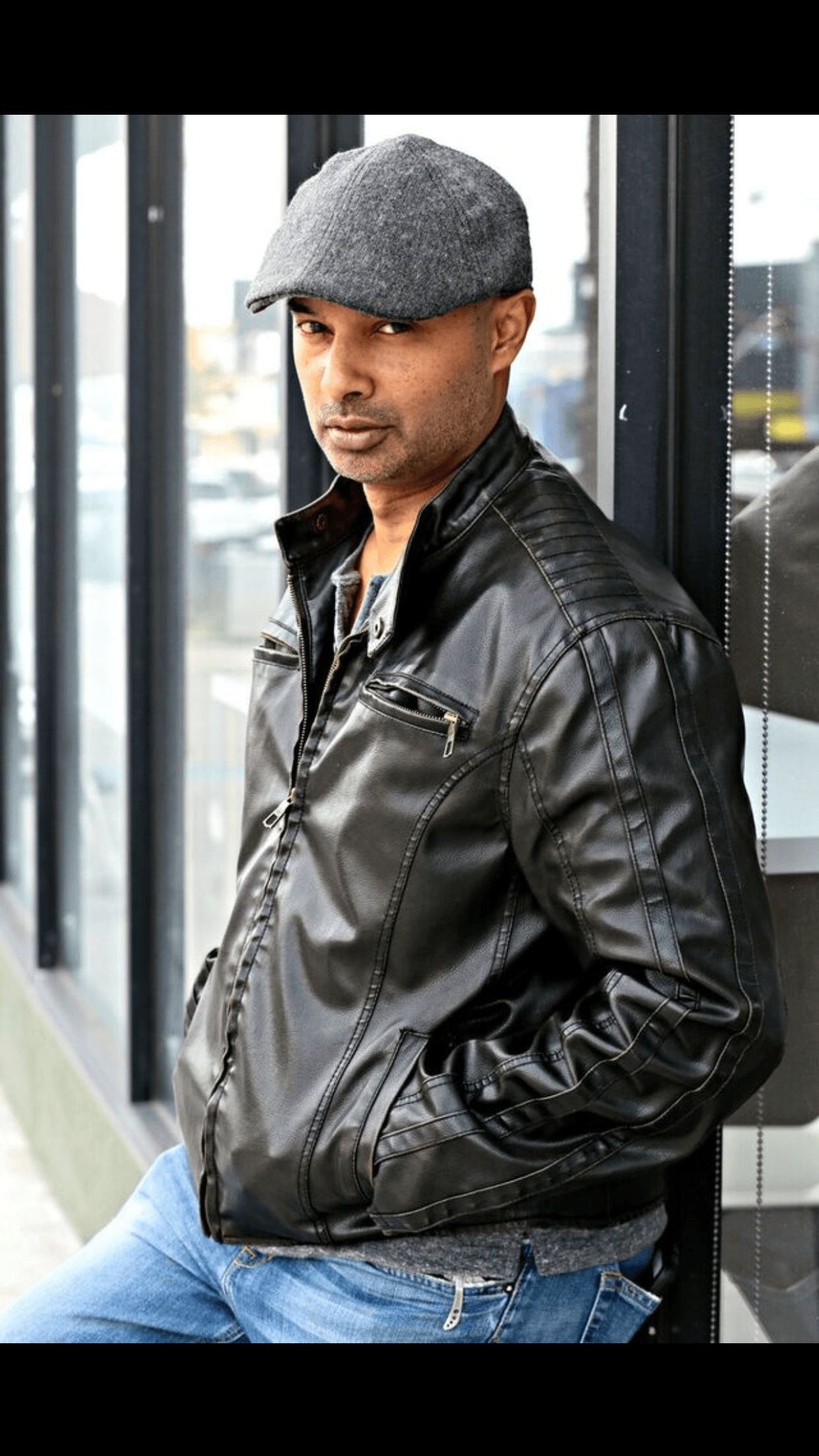 Man wearing a gray flat cap and black leather jacket standing outdoors near glass windows.
