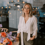 Woman with glasses and shoulder-length hair stands near a table with colorful flowers in a modern indoor setting.