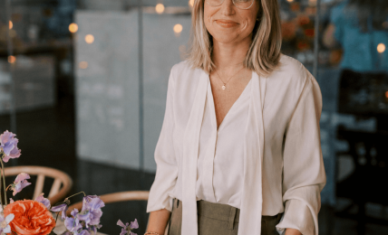 Woman with glasses and shoulder-length hair stands near a table with colorful flowers in a modern indoor setting.