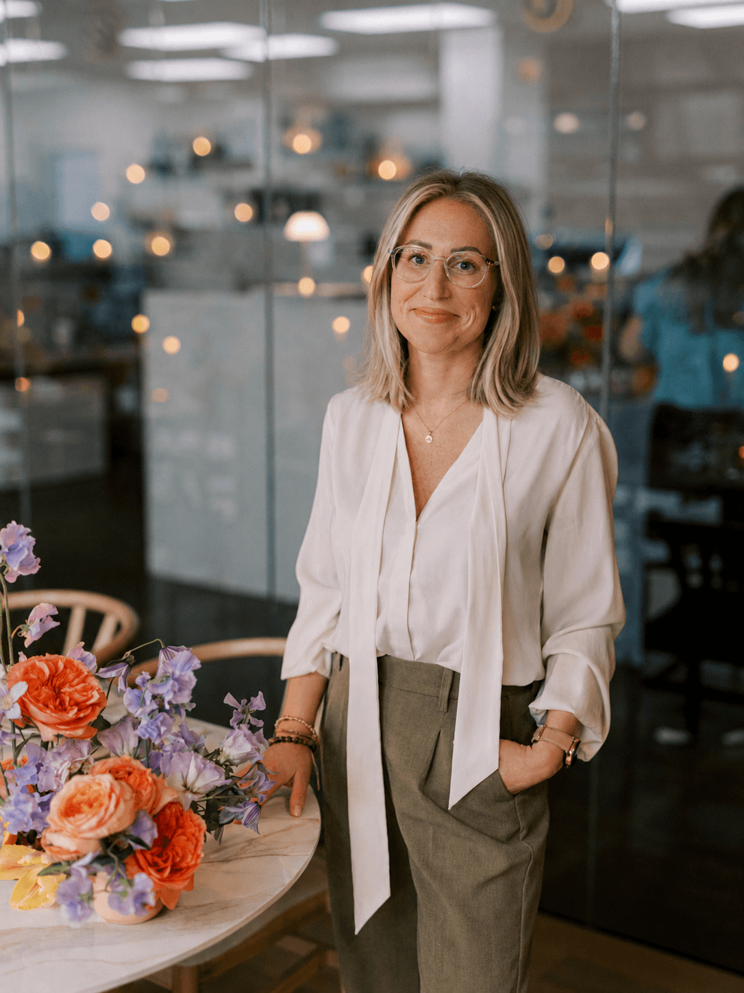 Woman with glasses and shoulder-length hair stands near a table with colorful flowers in a modern indoor setting.