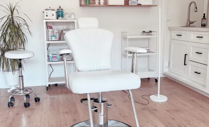 Medical or beauty treatment room with a white chair, shelves, and a lamp, on a wooden floor.