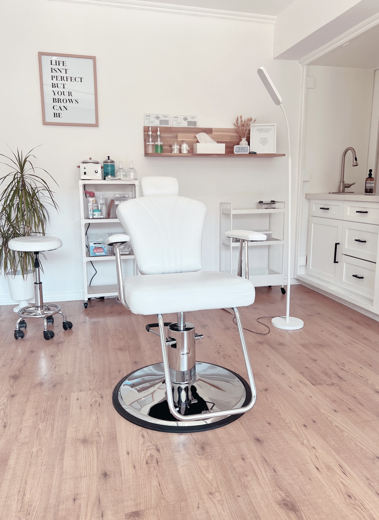 Medical or beauty treatment room with a white chair, shelves, and a lamp, on a wooden floor.