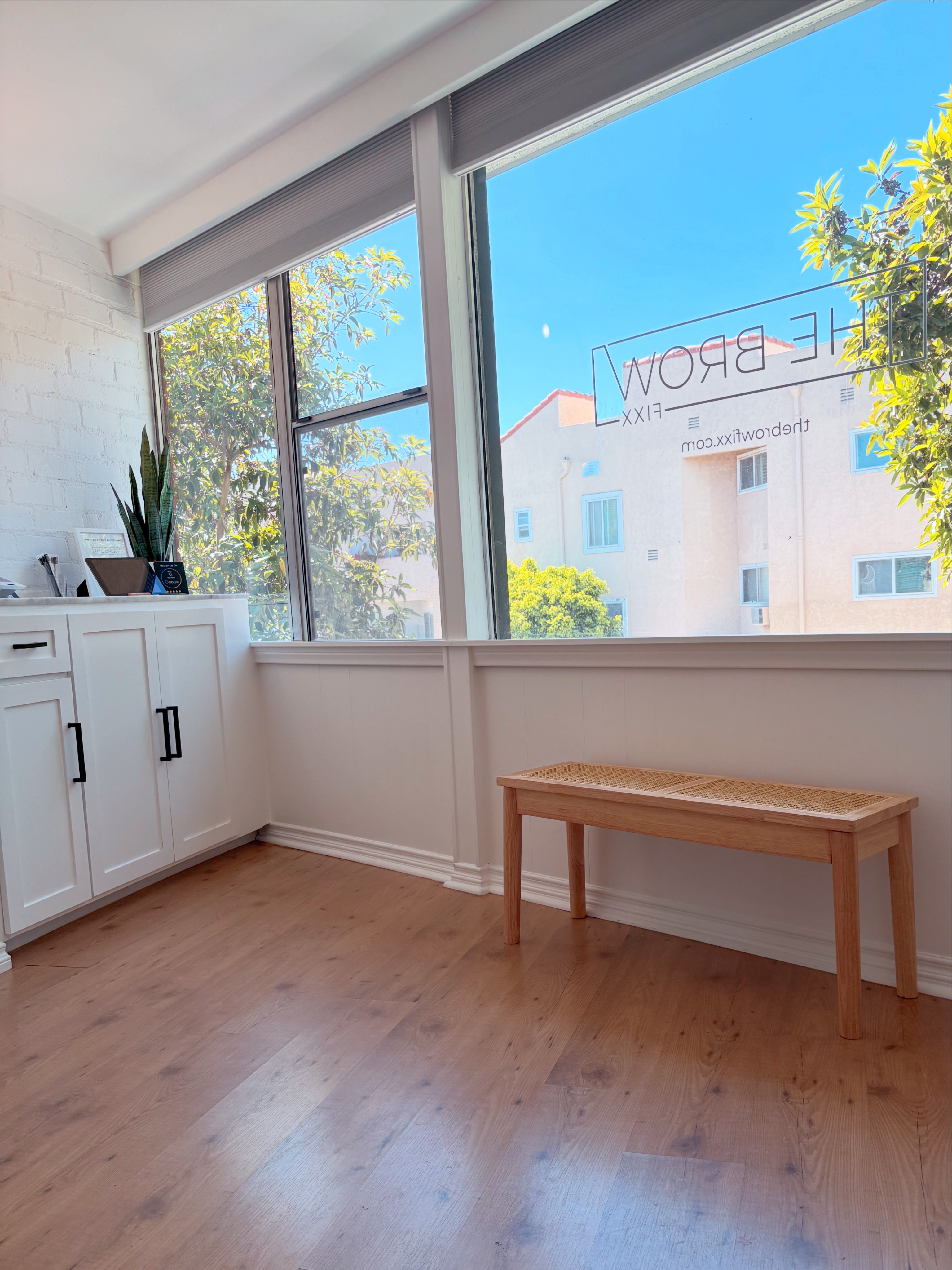 Room with large windows, white cabinet, potted plant, wooden bench, and view of building and trees outside.