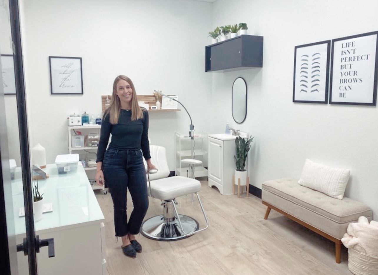 A woman stands in a modern, minimalistic room with white walls, a white chair, and framed artwork on the wall.