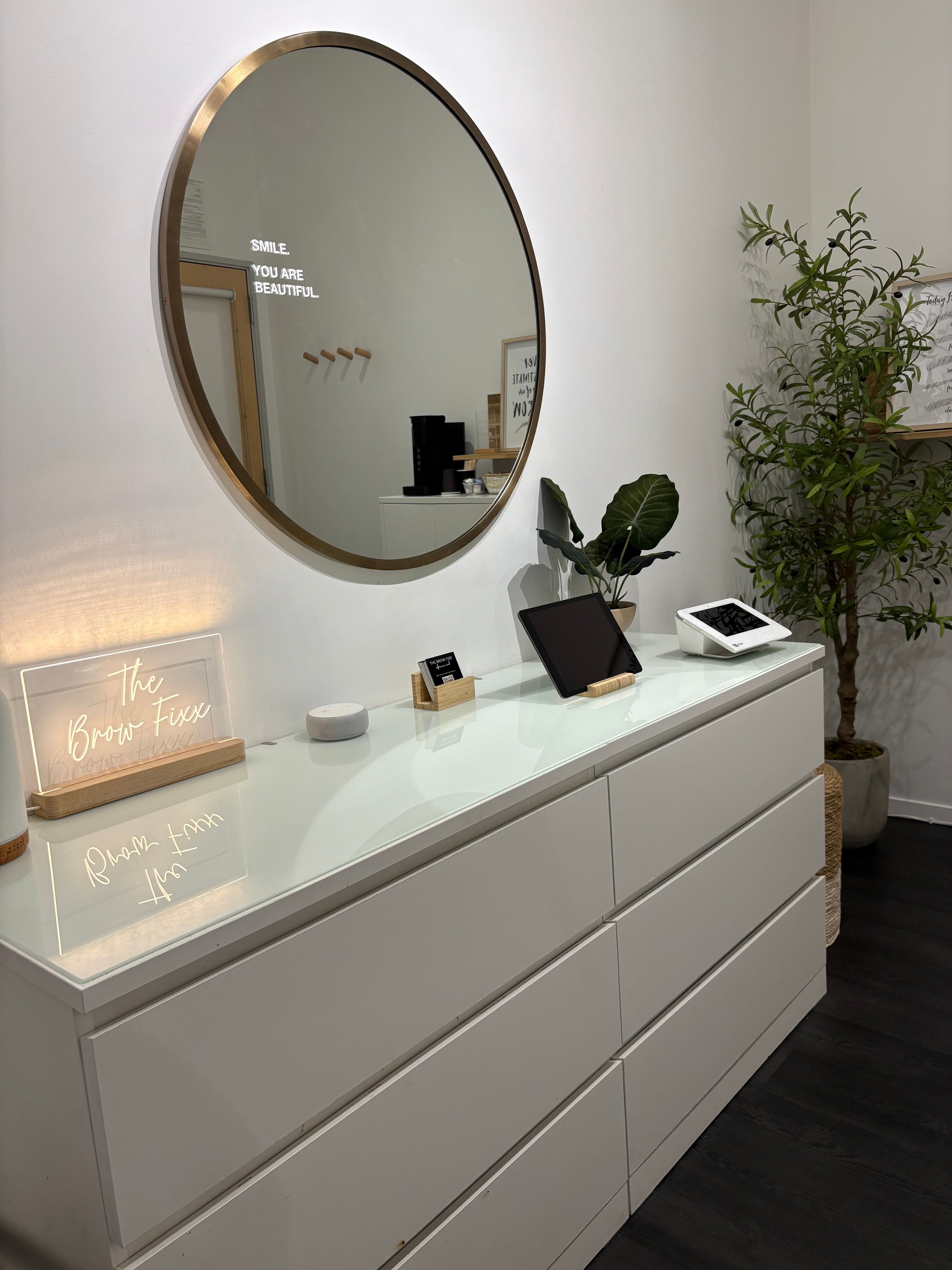 White dresser with a round mirror above, plants, a tablet, and decorative items on top, against a white wall.
