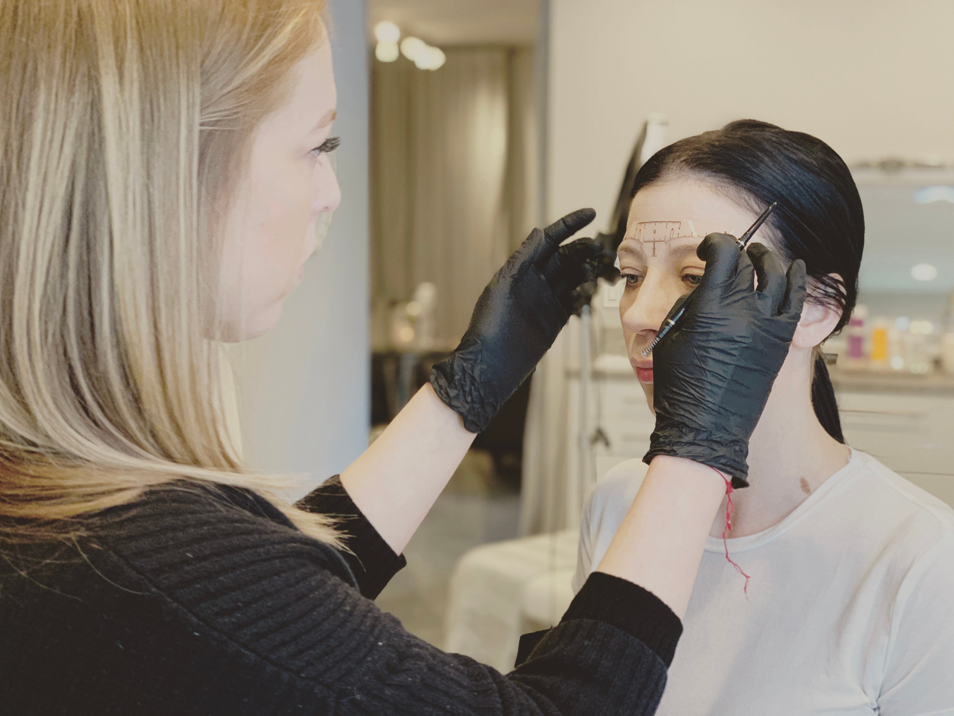 A woman with blonde hair performs a cosmetic procedure on another woman with dark hair, using gloves and tools.