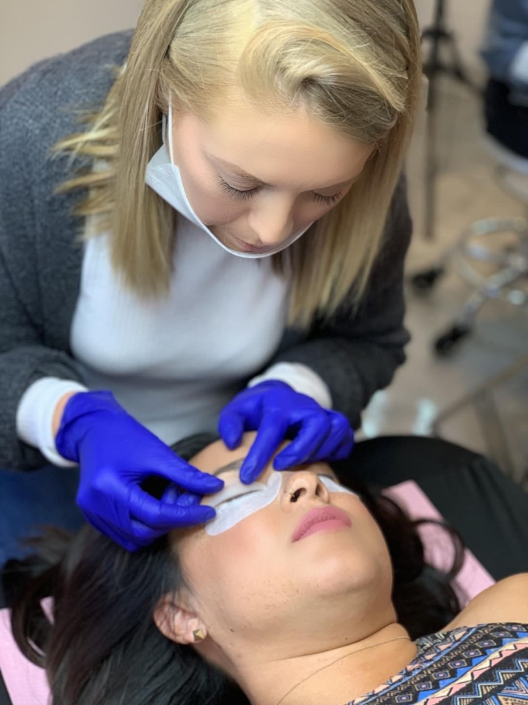 A woman with blonde hair and a face mask performs a procedure on a woman lying down, wearing blue gloves.
