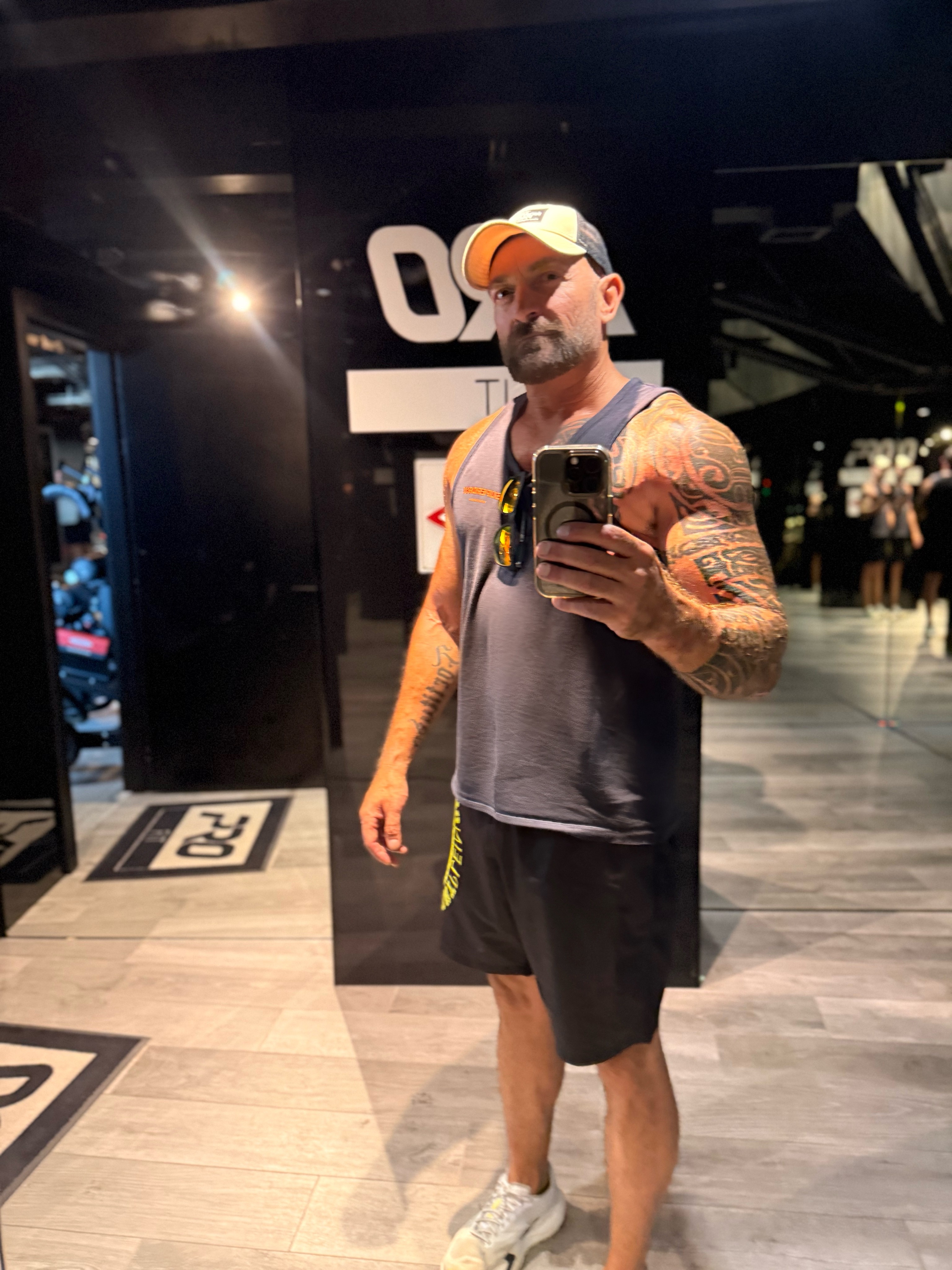 Man taking a mirror selfie in a gym, wearing a cap, sleeveless shirt, and shorts, with tattoos on arms.