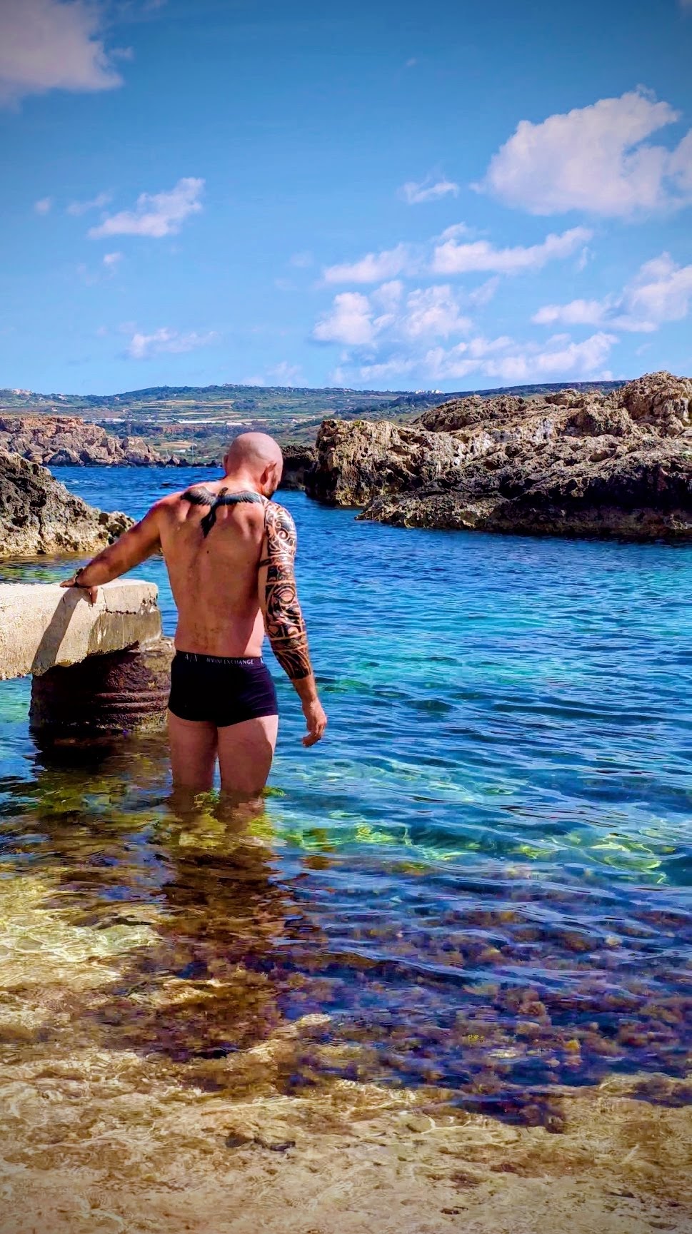 Man wades into clear blue water near rocky shoreline under a partly cloudy sky.