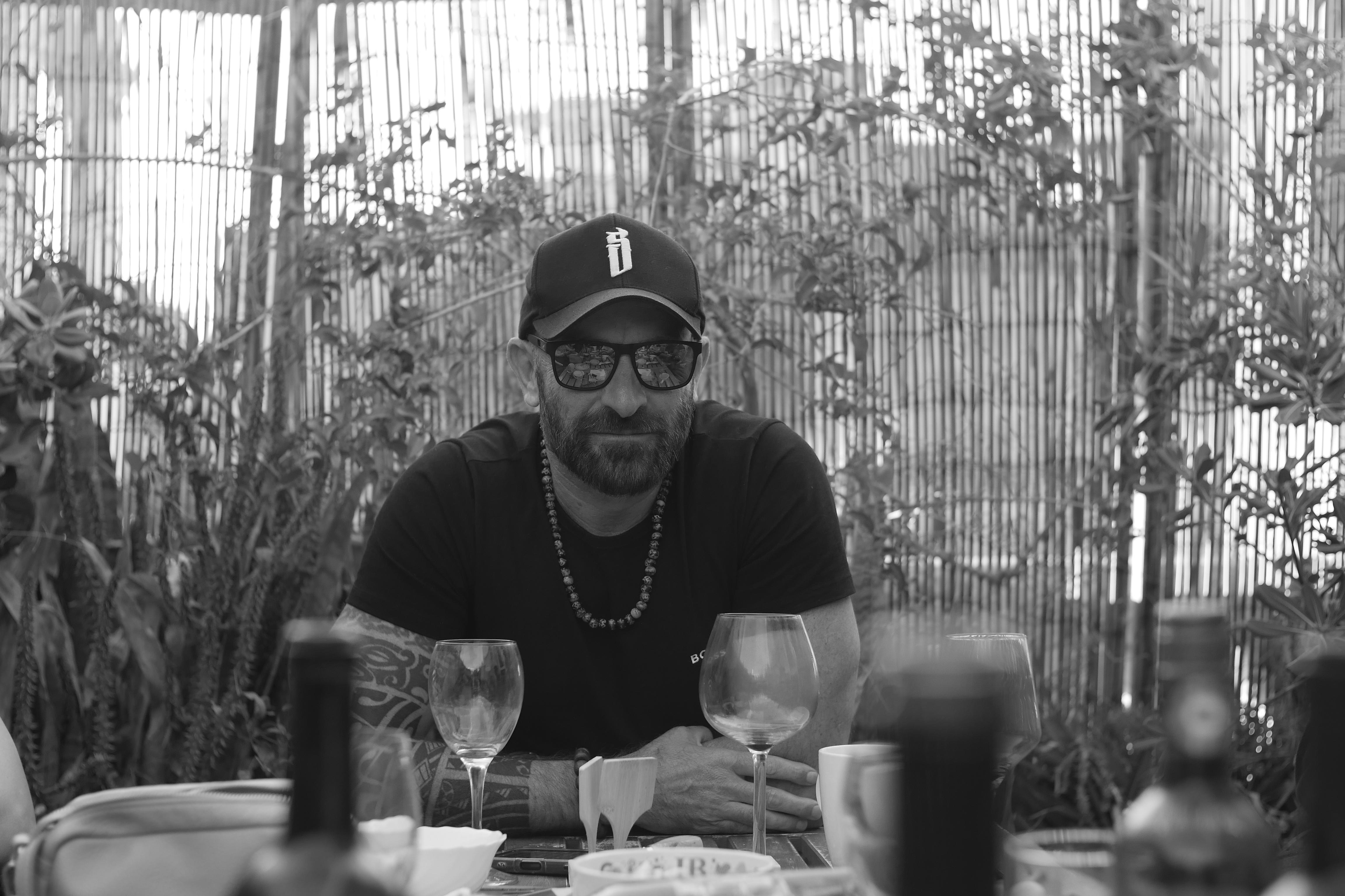 Man wearing glasses, cap, and chain sitting at a table with wine glasses and bottles outdoors.