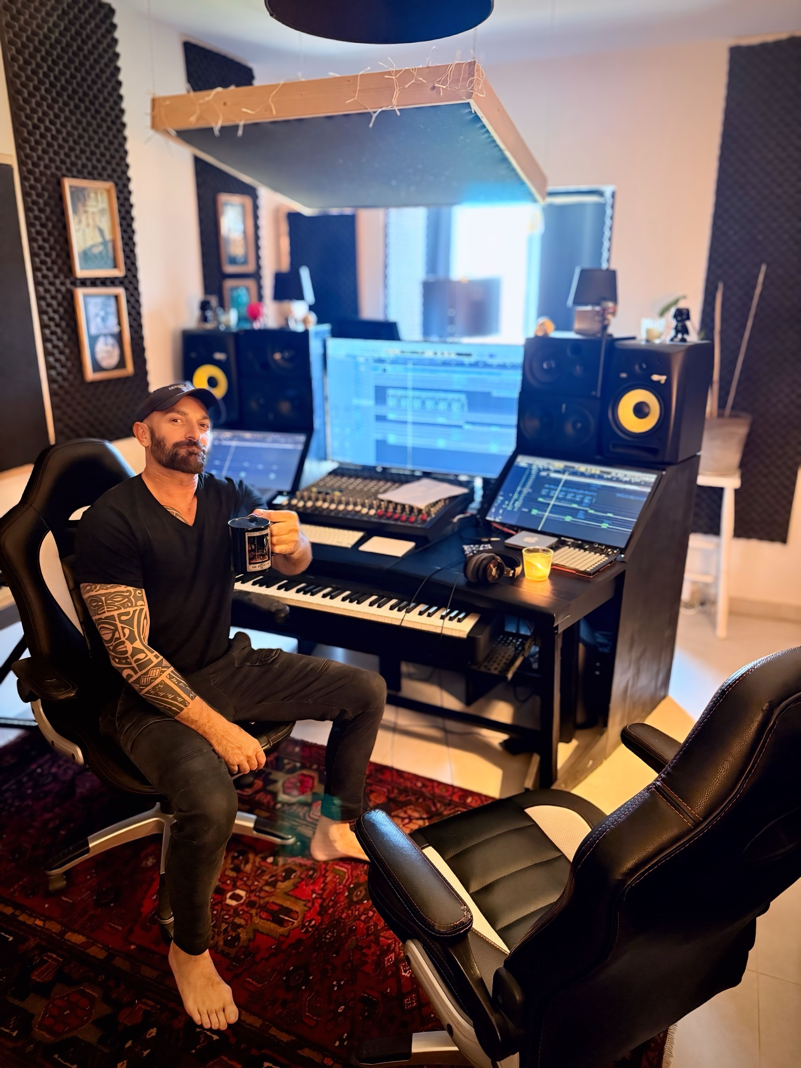 Man with tattoos sitting at a music production desk with computers and speakers in a studio.