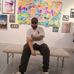 Person sitting on a bench in an art gallery with colorful paintings and framed pictures on the wall behind.