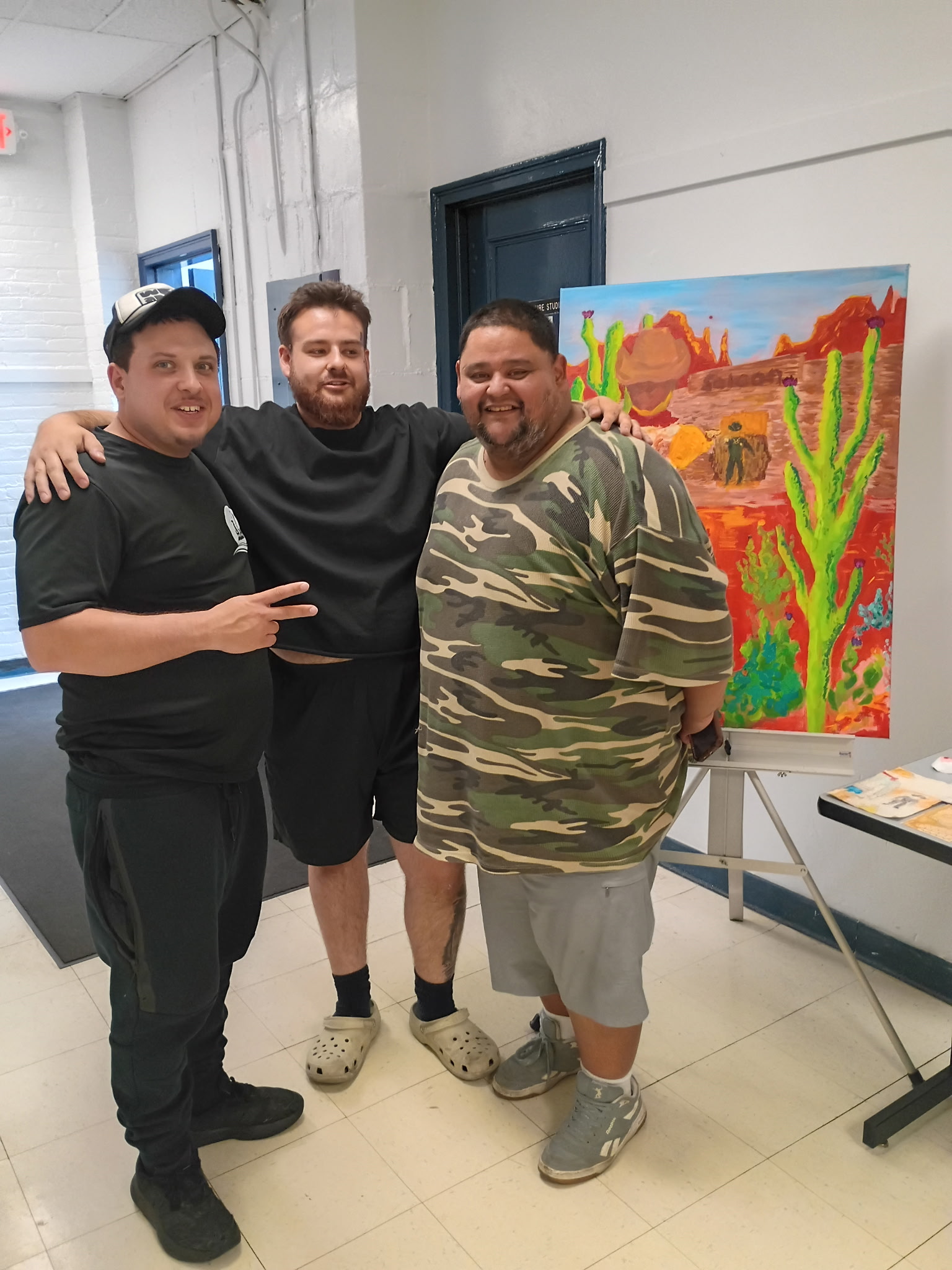 Three men standing indoors near a colorful desert landscape painting, smiling and posing together, one making a peace sign, casual clothing.